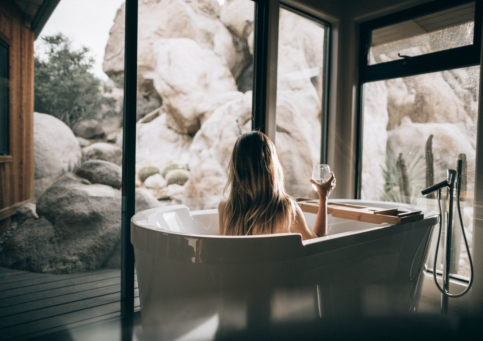 Image shows someone relaxing and unwinding in a large bathtub surrounded by glass and rocks outside.