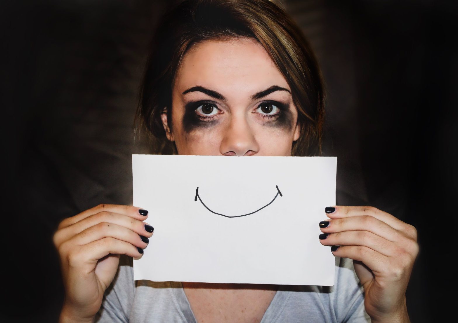 Image shows a person who looks like they've been crying but who is holding a piece of paper over their mouth with a smile drawn on it